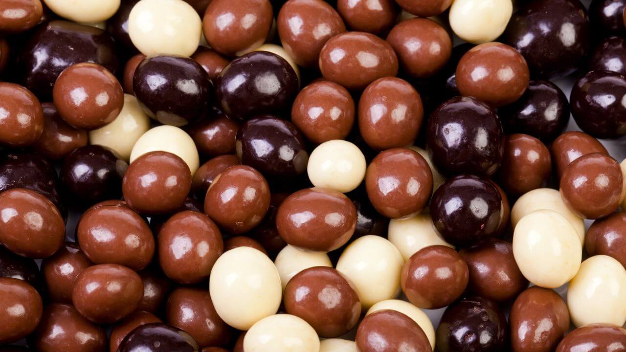 Como ganhar dinheiro com Drageados - Um grupo de confeitos de chocolate redondos e polidos, conhecidos como drageados, preenche todo o quadro da imagem. Eles variam em cores e acabamentos, incluindo tons de chocolate amargo (marrom escuro), chocolate ao leite (marrom médio) e chocolate branco (creme). Os doces possuem uma superfície lisa e brilhante que reflete a luz, criando pequenos pontos de realce em cada unidade.