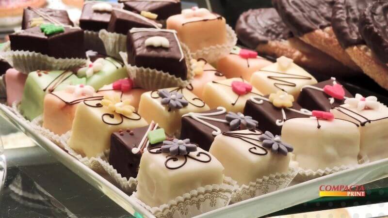 Petit Fours Embalagem Uma bandeja branca repleta de mini bolos decorados (petits fours) dispostos em forminhas de papel plissado branco. Os doces possuem formatos quadrados e retangulares, cobertos com glacê ou chocolate em tons de marrom escuro, creme, rosa claro e verde menta. Cada unidade é finalizada com detalhes delicados, como pequenas flores e corações de açúcar coloridos, além de finos riscos decorativos de calda. Ao fundo, à direita, é possível ver uma pilha de biscoitos grandes cobertos de chocolate. No canto inferior direito, há um logotipo com o texto "COMPACTA PRINT".