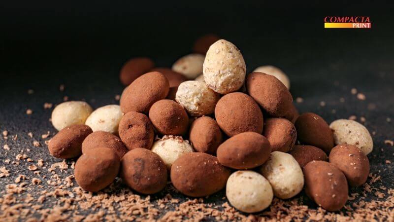 Produção de drageados Fotografia de uma pequena pilha de drageados com acabamento rústico sobre uma superfície escura. A composição mistura drageados marrons escuros, com aspecto fosco por estarem polvilhados com cacau em pó, e drageados de cor clara com textura irregular. Pequenas raspas de chocolate estão espalhadas na base ao redor dos doces. No canto superior direito, encontra-se o logotipo da Compacta Print.