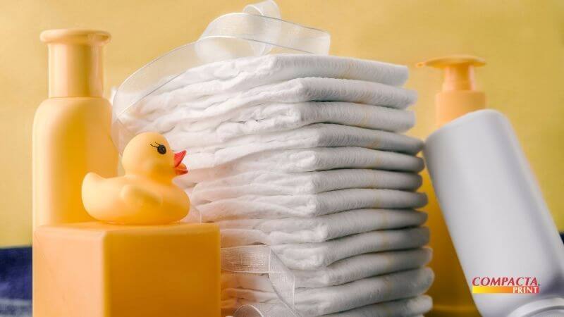 Fabricar em casa garante - Fraldas Uma pilha alta de fraldas brancas descartáveis organizadas verticalmente. Ao lado, há produtos de higiene infantil em embalagens amarelas e brancas, além de um patinho de borracha amarelo sobre um bloco. O fundo é amarelo claro, e o logotipo "Compacta Print" aparece no canto inferior direito.