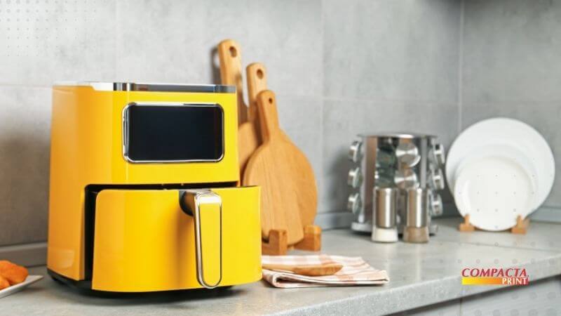 Por que a Air Fryer Mudou o Jogo? Uma air fryer de design moderno na cor amarela posicionada sobre uma bancada de cozinha cinza. Ao fundo, veem-se tábuas de carne de madeira, pratos brancos e um porta-condimentos. No canto inferior direito, há o logotipo "Compacta Print".