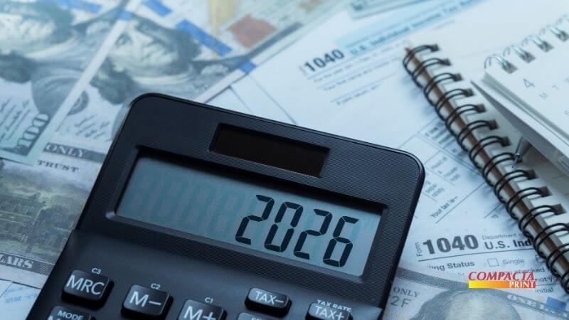Empreender 2026 Close-up de uma calculadora preta onde o visor exibe o número "2026". A calculadora está sobreposta a notas de cem dólares e formulários de impostos (Form 1040). Ao lado, vê-se parte de um caderno de espiral e uma caneta. O logotipo "Compacta Print" está posicionado no canto inferior direito.