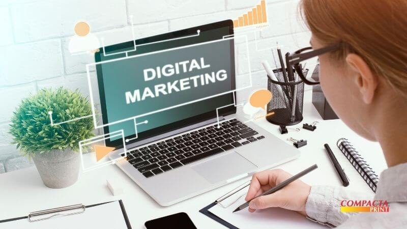 Marketing de baixo custo Uma pessoa de cabelos ruivos e óculos está sentada em uma mesa de trabalho branca, olhando para a tela de um notebook que exibe a mensagem "DIGITAL MARKETING" em destaque. Gráficos de ícones flutuantes, como um perfil de usuário, gráficos de barras e balões de conversa, cercam o notebook. Ela está escrevendo em um papel com uma caneta. Na mesa, há uma planta em um vaso de cimento, um organizador de material de escritório com canetas e clipes, e um smartphone. O logotipo da Compacta Print está no canto inferior direito.
