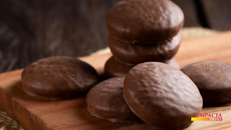 Engenharia de Cardápio e Precificação Fotografia em close-up de pães de mel cobertos com chocolate. Alguns estão empilhados ao fundo e outros em destaque no primeiro plano sobre uma base de madeira. No canto inferior direito, há o logotipo "Compacta Print".